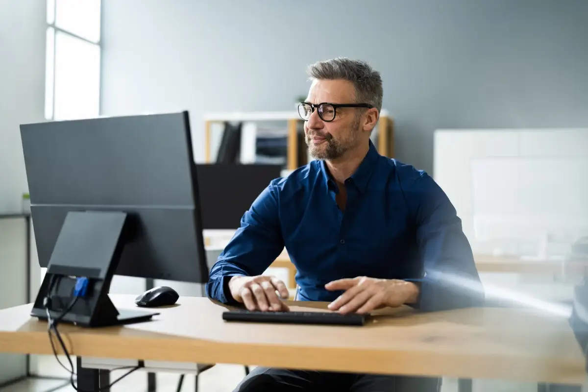 Homem sentado em escritório, com óculos e camisa azul, postura relaxada enquanto trabalha em seu computador.